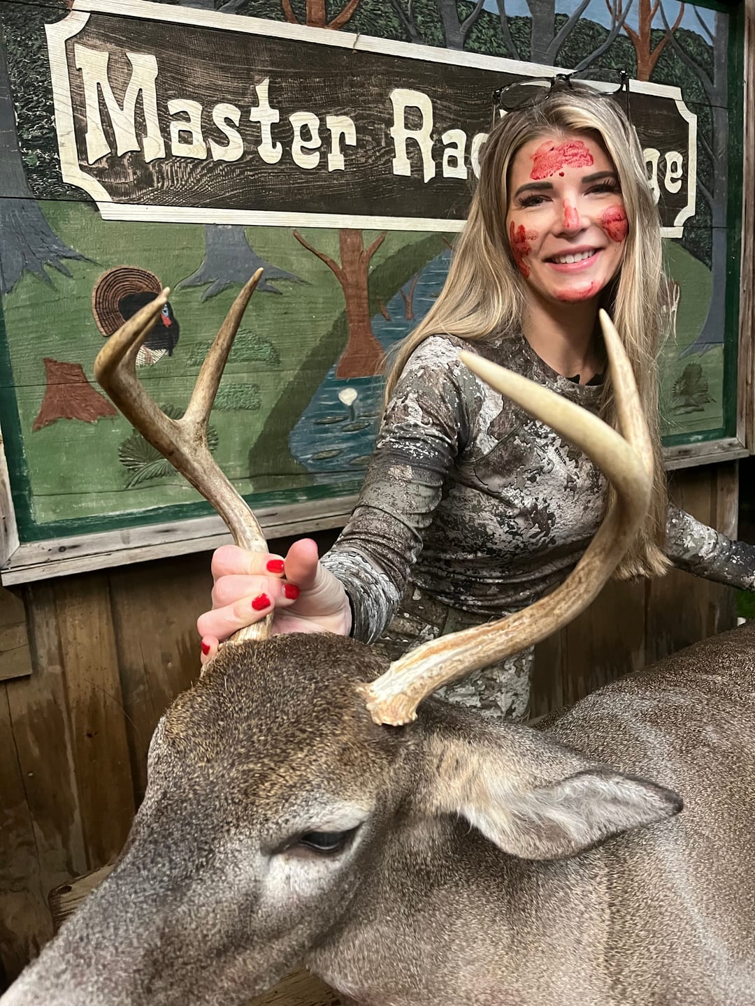 A woman with blood on her face poses with a harvested whitetail deer in front of a "Master Rack Lodge" sign.