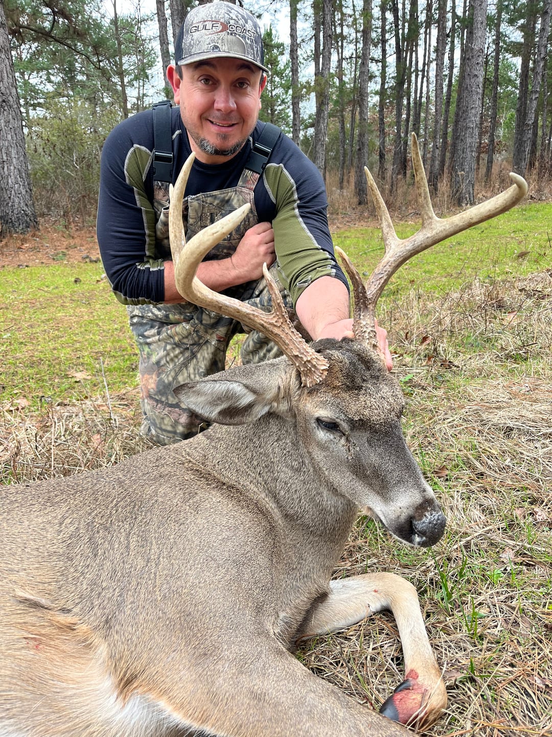 A hunter in camouflage poses with a harvested whitetail deer in a wooded area.