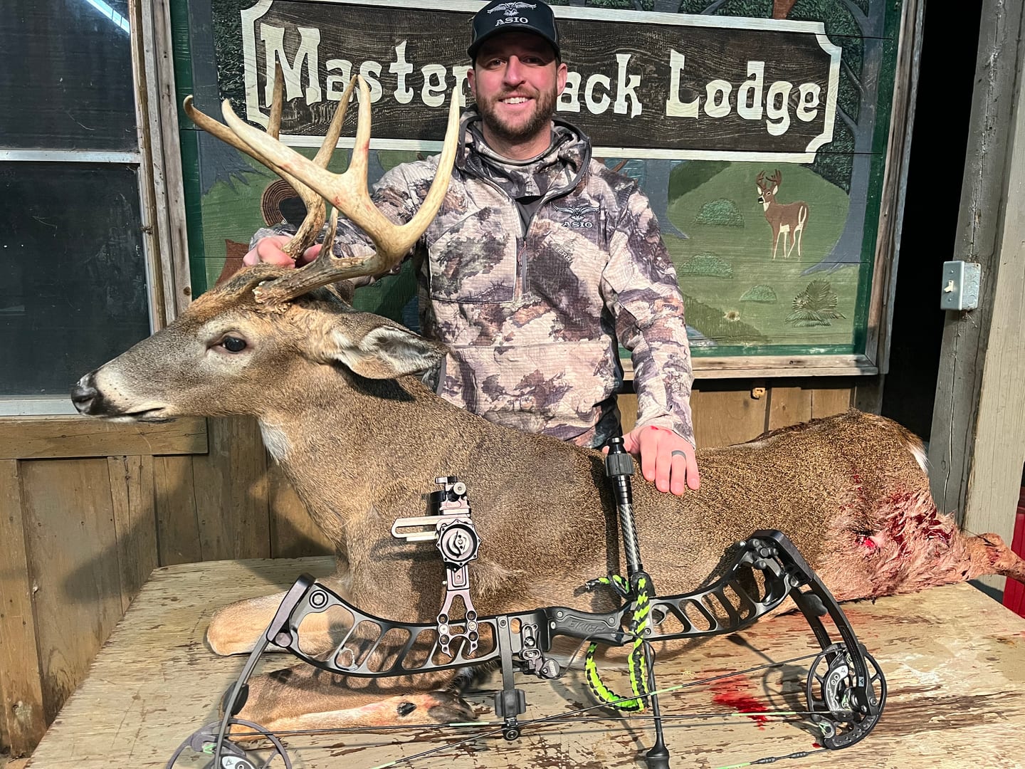 A man in camouflage poses with a harvested whitetail deer and a bow in front of a "Master Rack Lodge" sign.