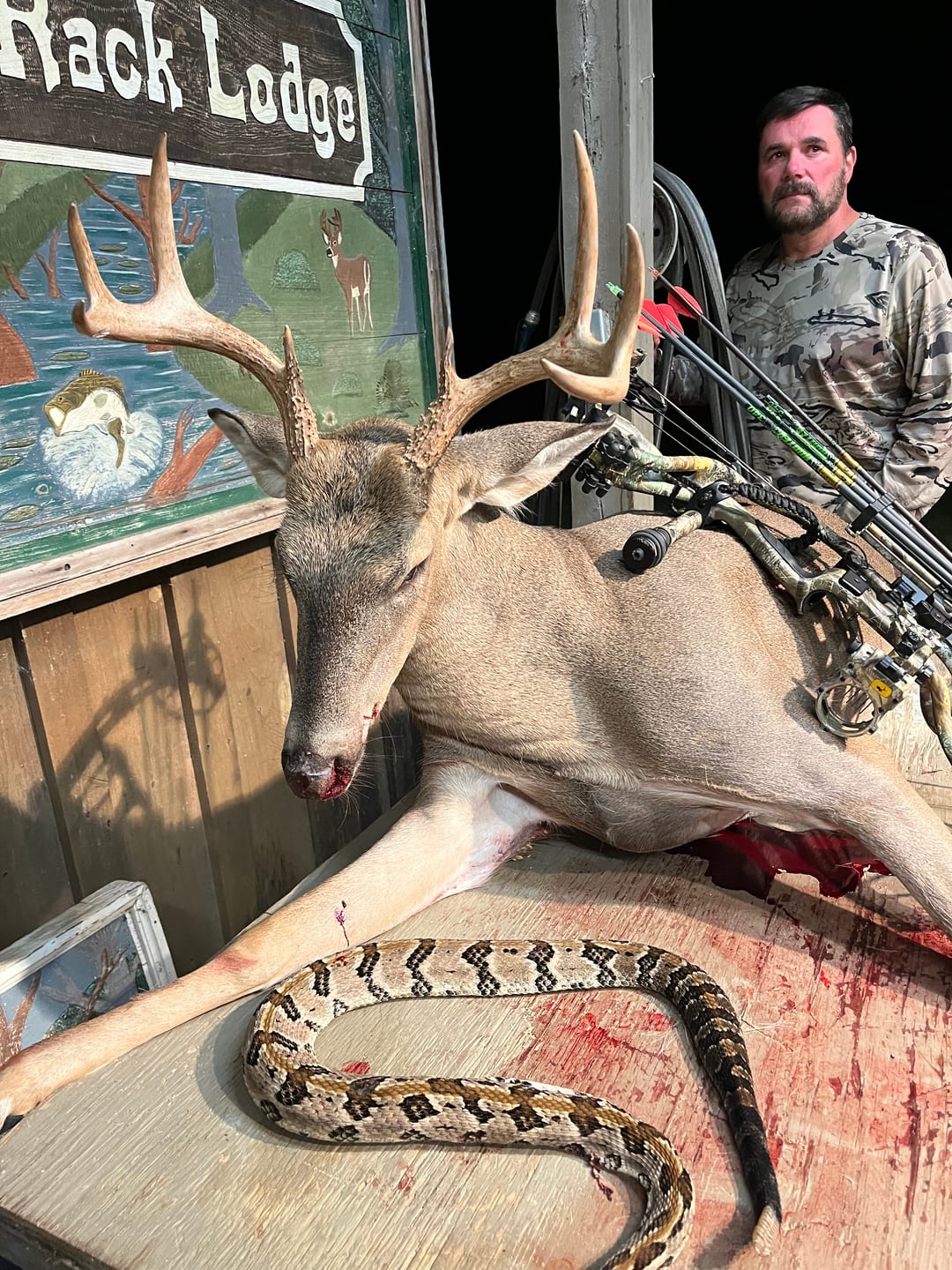 A hunter poses with a harvested whitetail deer and a snake on a table.