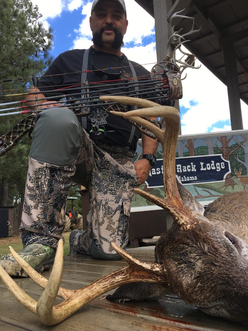 A man in camouflage kneels with a harvested whitetail deer and a bow on a wooden deck, with a "Master Rack Lodge" sign in the background.