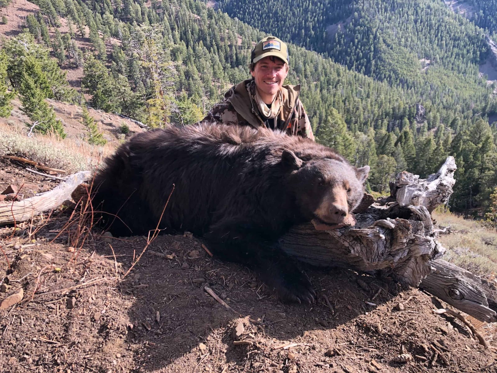 A hunter poses with a harvested black bear on a hillside with a forested mountain in the background.