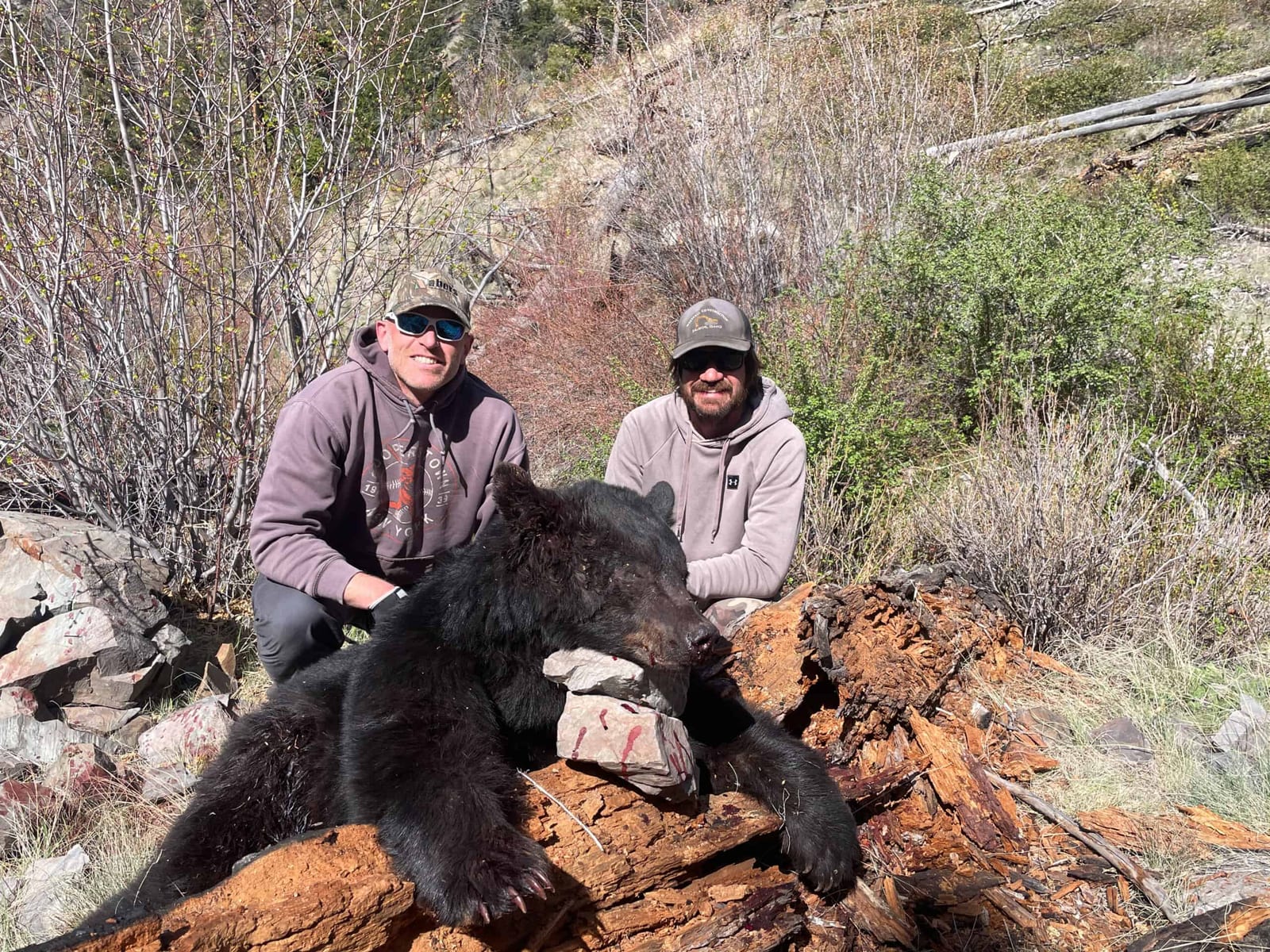 Two hunters pose with a harvested black bear on a rocky, brushy hillside.