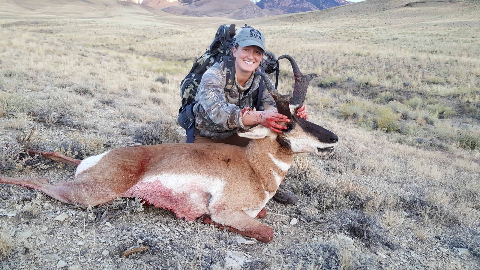 A woman in hunting gear poses with a harvested pronghorn in a dry, grassy field.