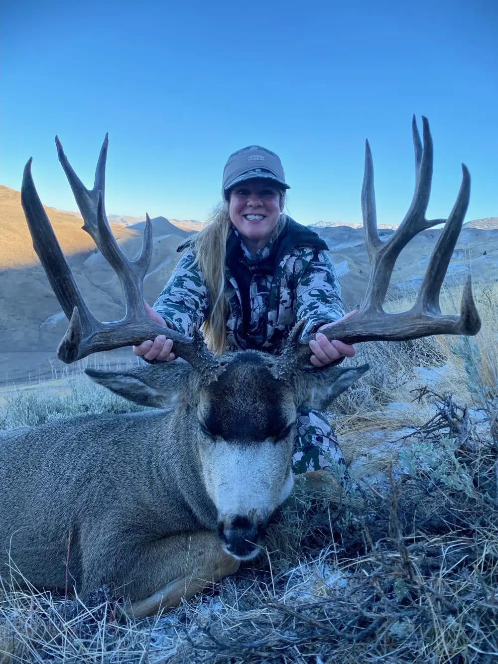 A woman in camouflage poses with a harvested mule deer in a mountainous landscape.