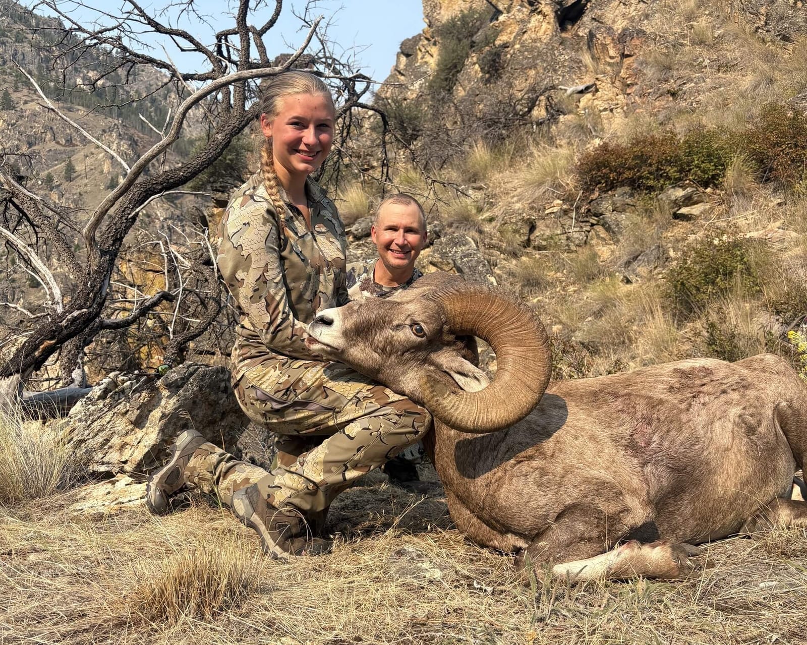 A young woman and a man pose with a harvested bighorn sheep in a rocky, mountainous landscape.