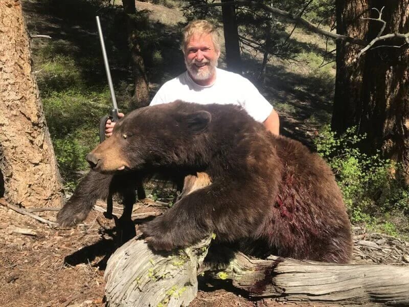 A hunter poses with a harvested black bear in a forest.