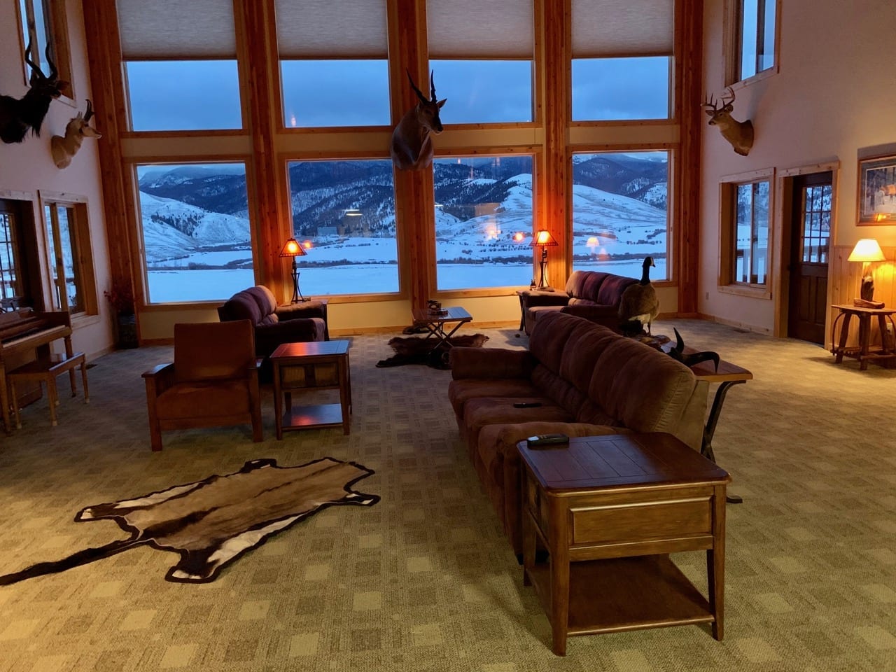 A spacious lodge interior with large windows overlooking a snowy mountain landscape, featuring multiple taxidermy mounts and a fur rug.