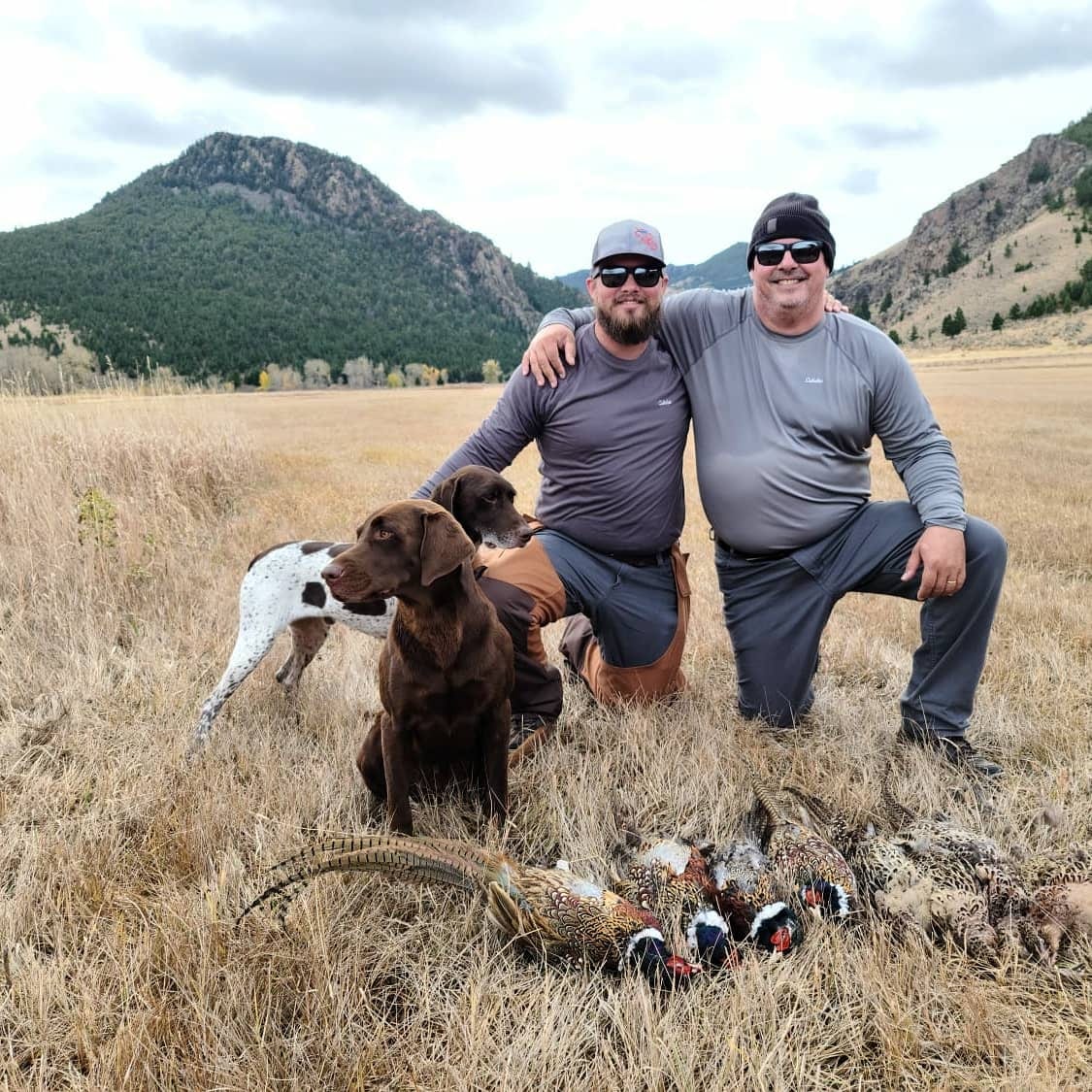 Two hunters pose with their dogs and harvested ring-necked pheasants in a field with mountains in the background.