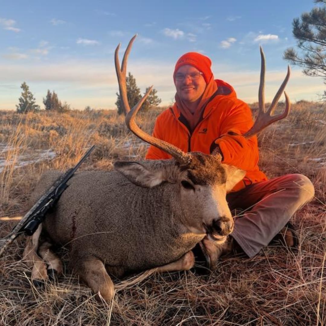 A hunter in an orange hat and jacket poses with a harvested mule deer in a grassy field.