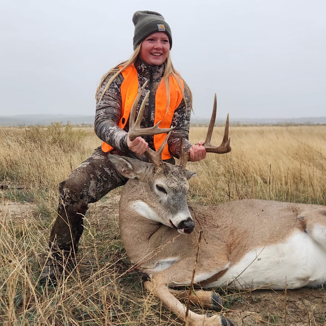 A young woman in hunting attire poses with a harvested whitetail deer in a grassy field.