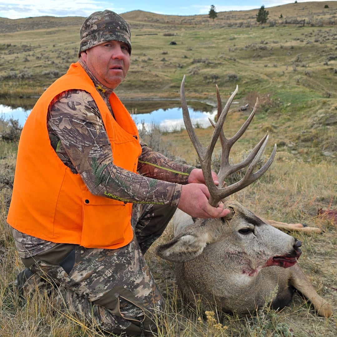 A hunter kneels next to a harvested mule deer in a grassy field with a pond in the background.
