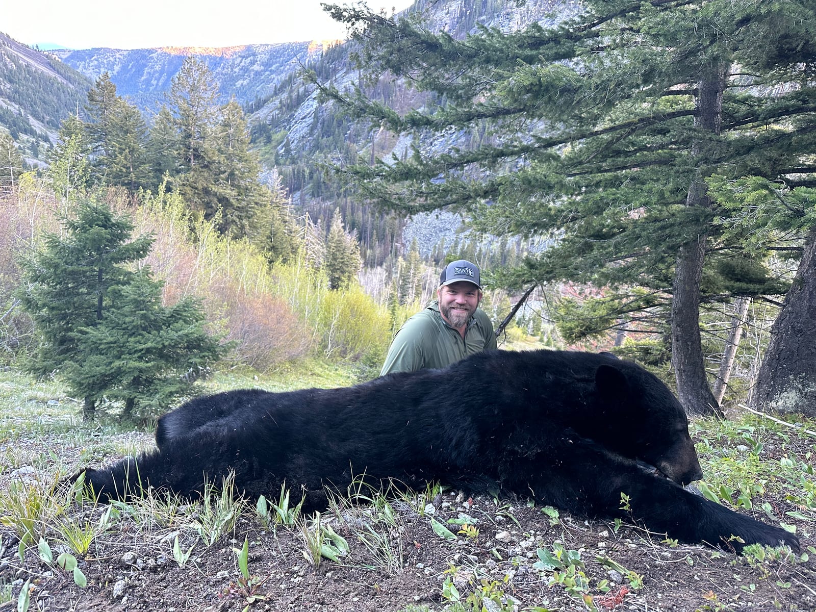 A hunter poses with a harvested black bear in a mountainous, forested landscape.