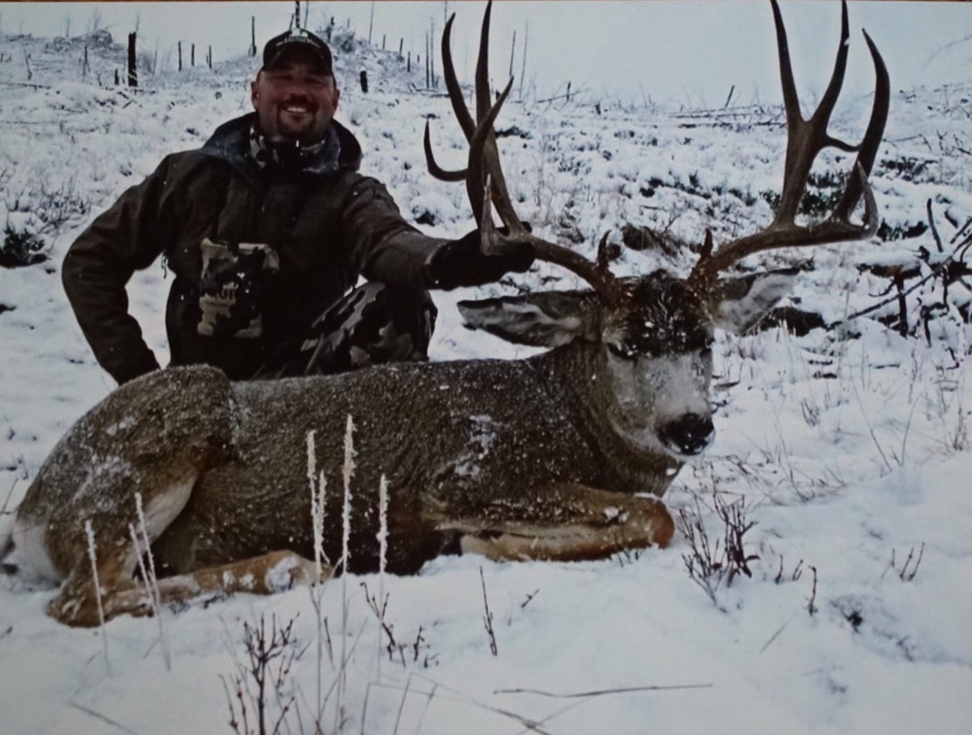 A hunter poses with a harvested mule deer in a snowy landscape.