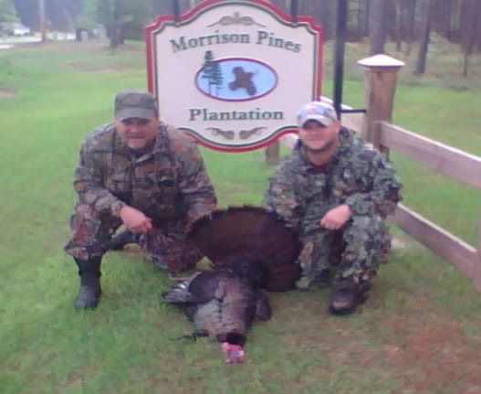 Two hunters in camouflage pose with a harvested wild turkey in front of a sign for Morrison Pines Plantation.
