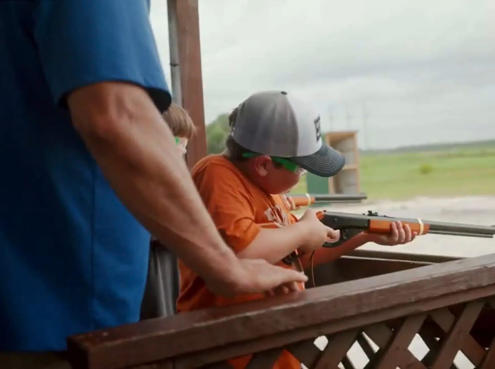 A child in a cap and safety glasses holds a rifle at a shooting range, with an adult's arm visible in the foreground.