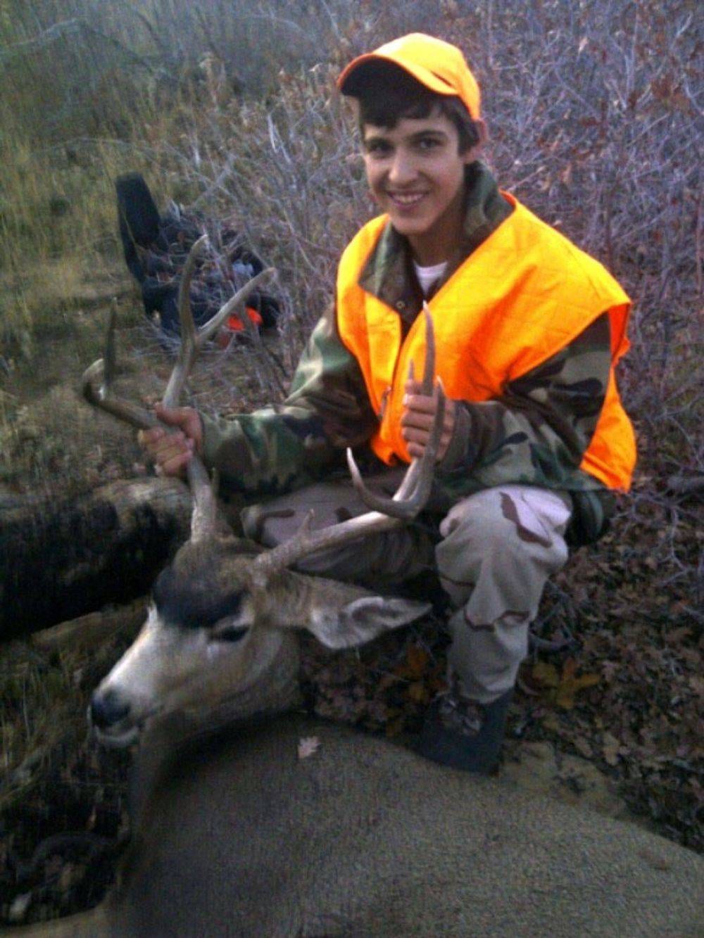A young hunter in an orange vest poses with a harvested mule deer.