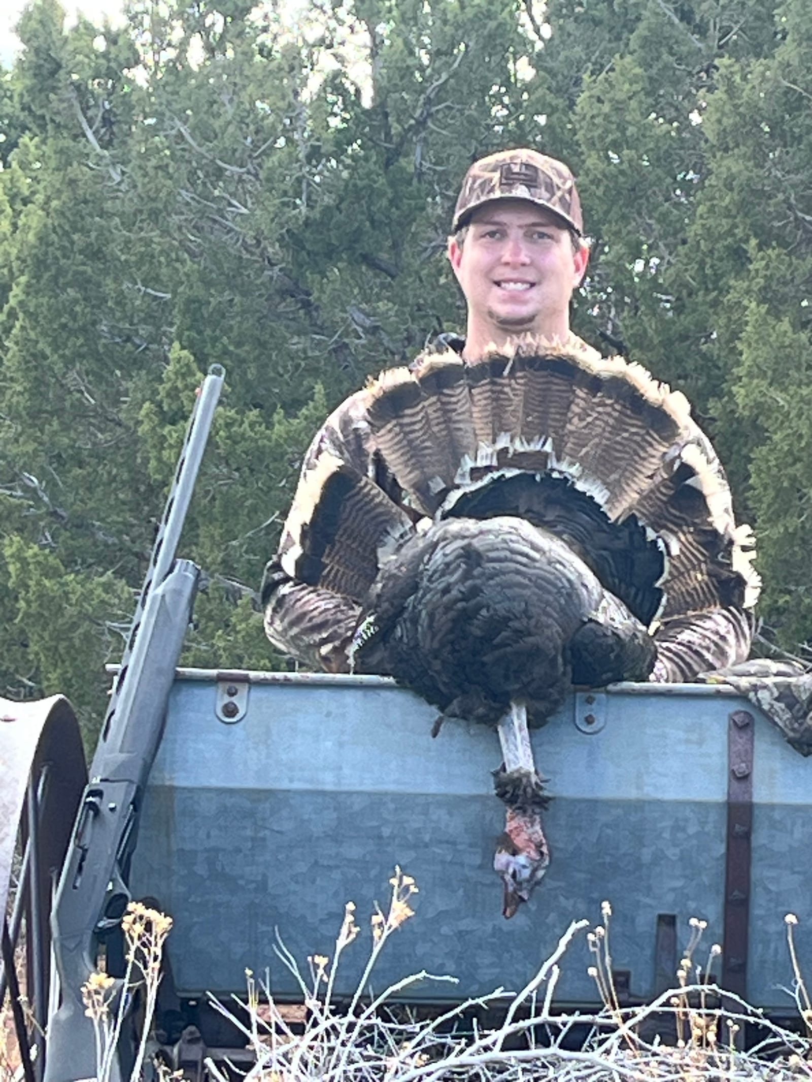 A hunter in camouflage poses with a harvested wild turkey on the back of a vehicle.
