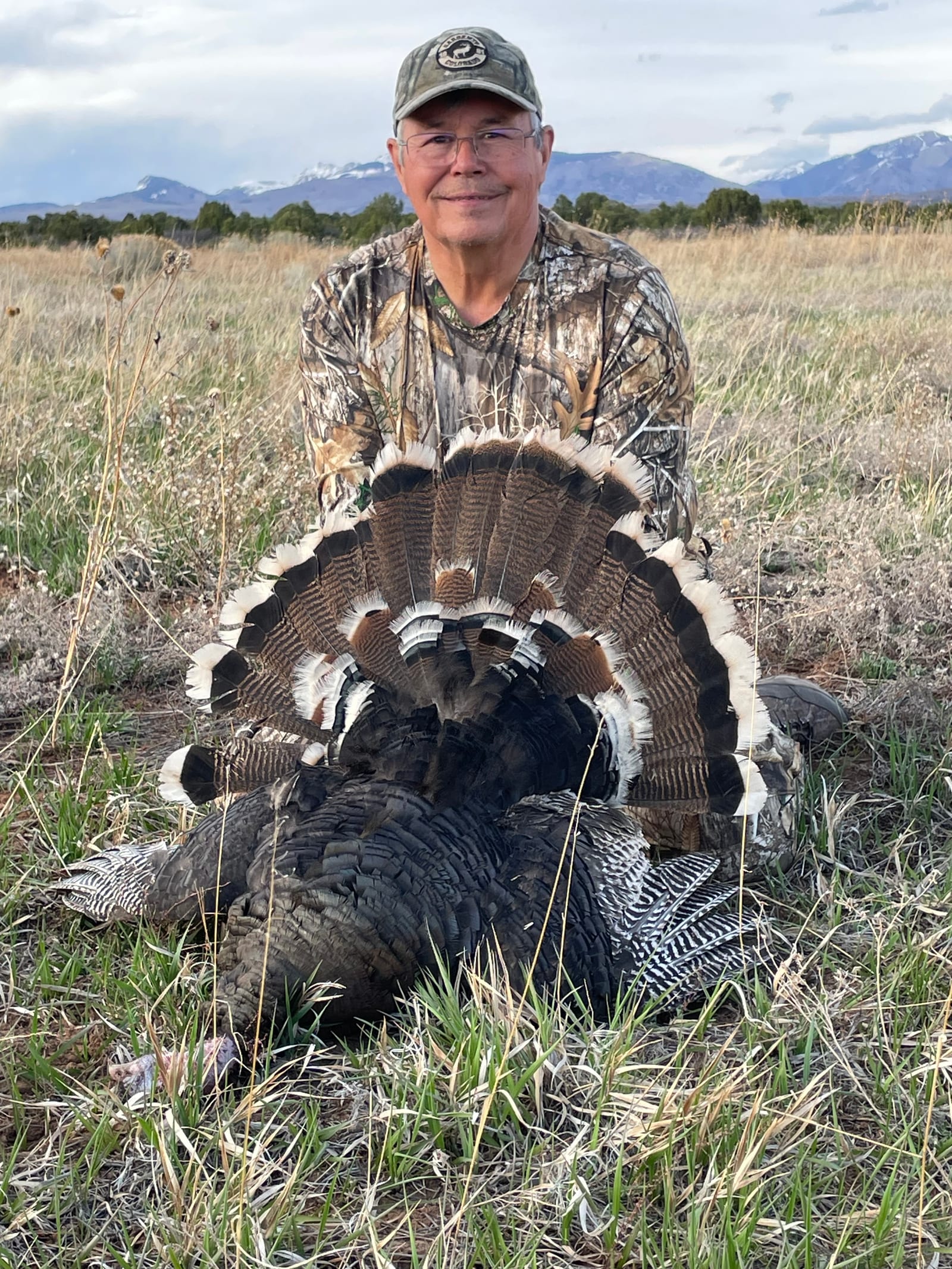 A man in camouflage poses with a harvested wild turkey in a grassy field with mountains in the background.