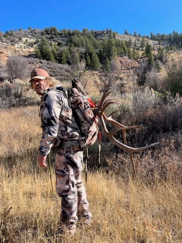 A hunter carries an elk antler on his back in a mountainous, grassy field.
