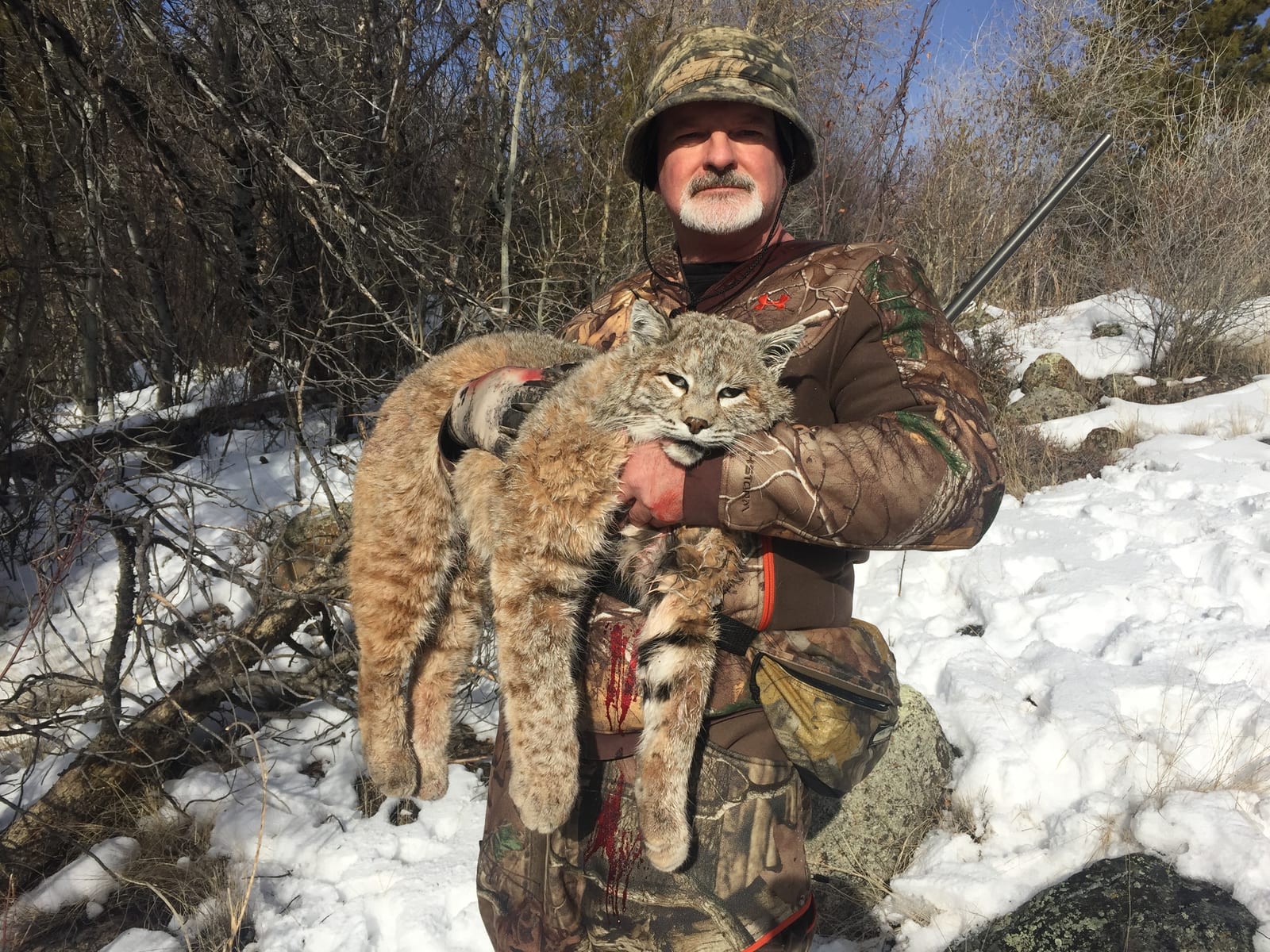 A hunter in camouflage holds a harvested bobcat in a snowy, wooded environment.