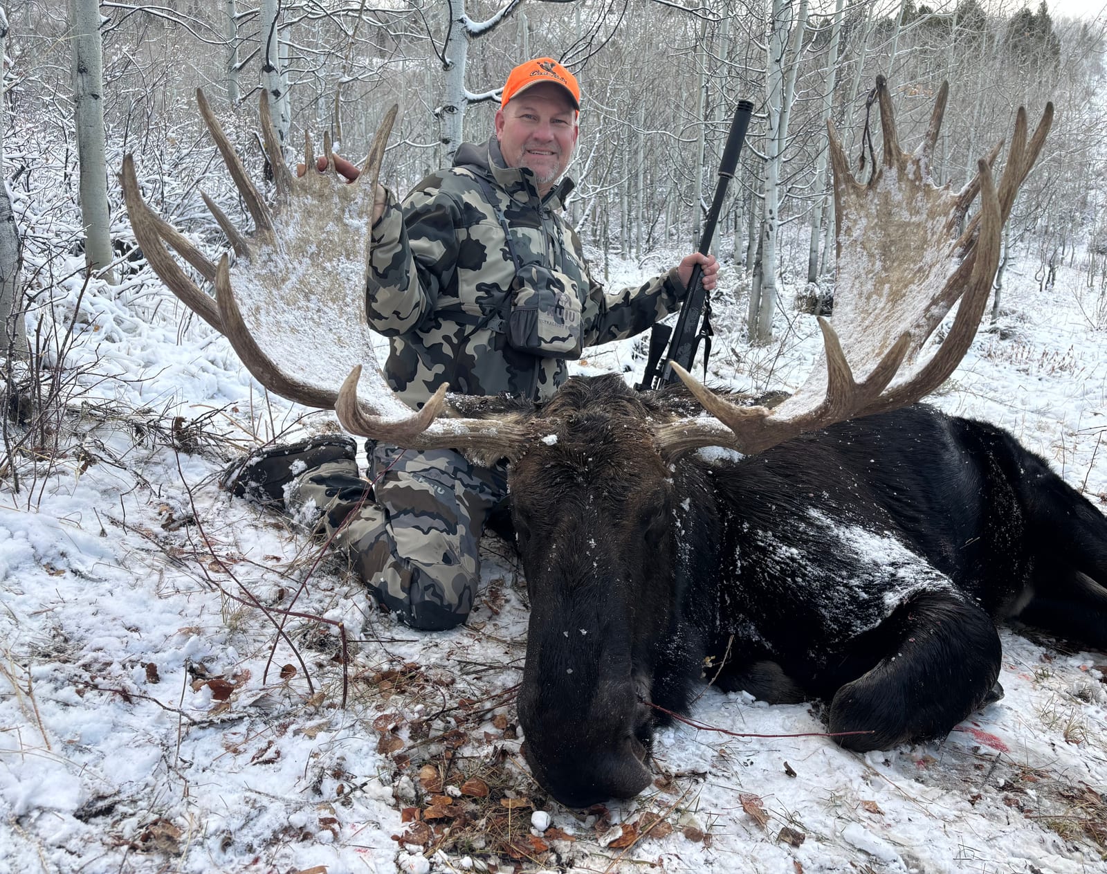 A man in camouflage and an orange hat poses with a large harvested moose in a snowy, wooded area.
