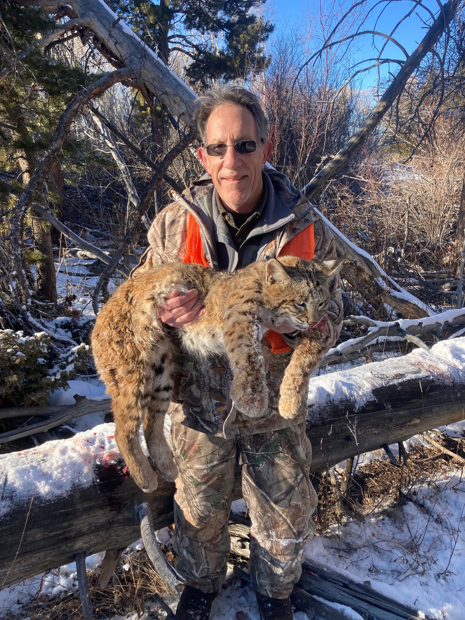 A hunter in camouflage clothing holds a harvested bobcat in a snowy, wooded area.