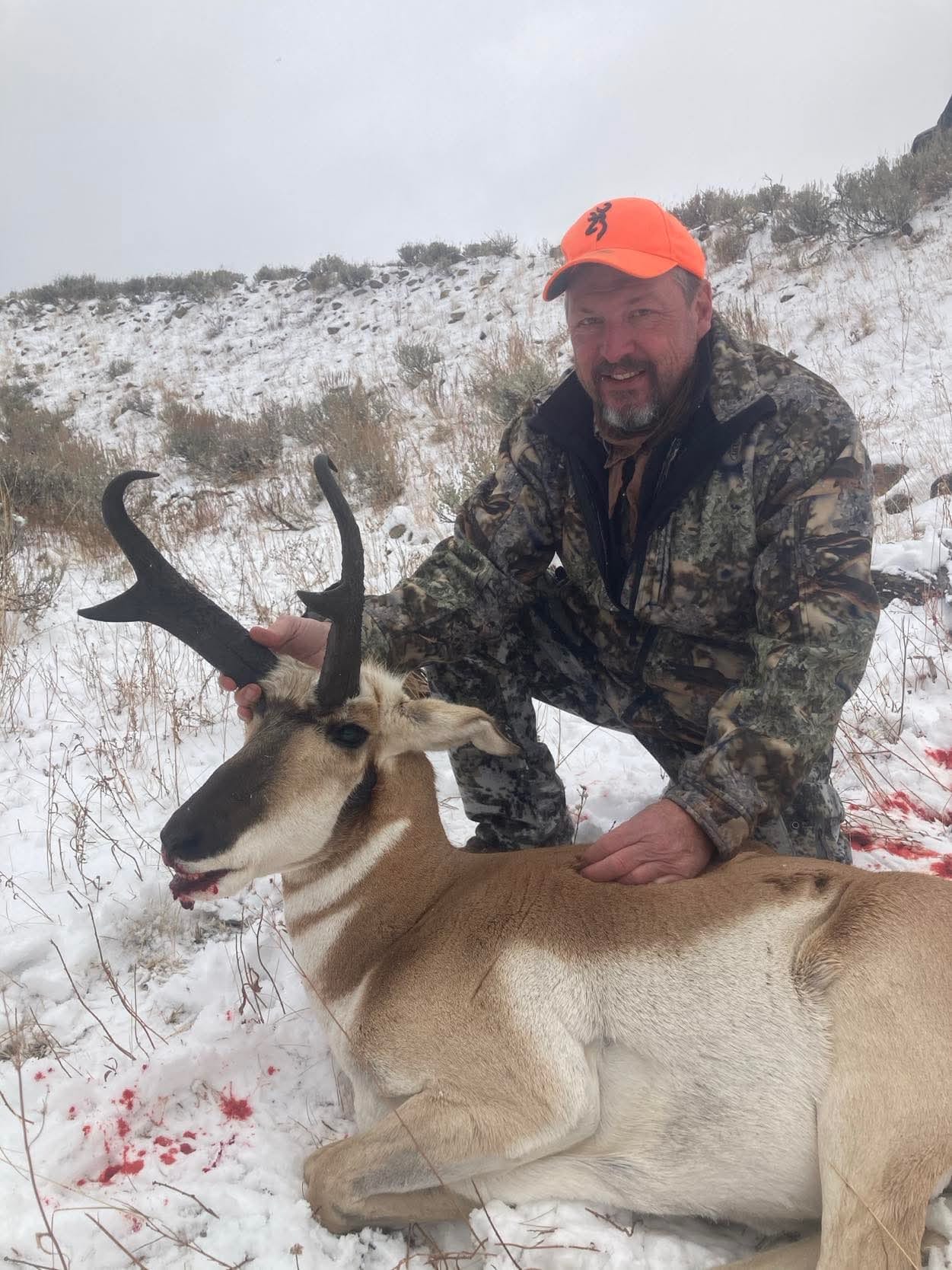 A man in camouflage and an orange hat poses with a harvested pronghorn in a snowy field.