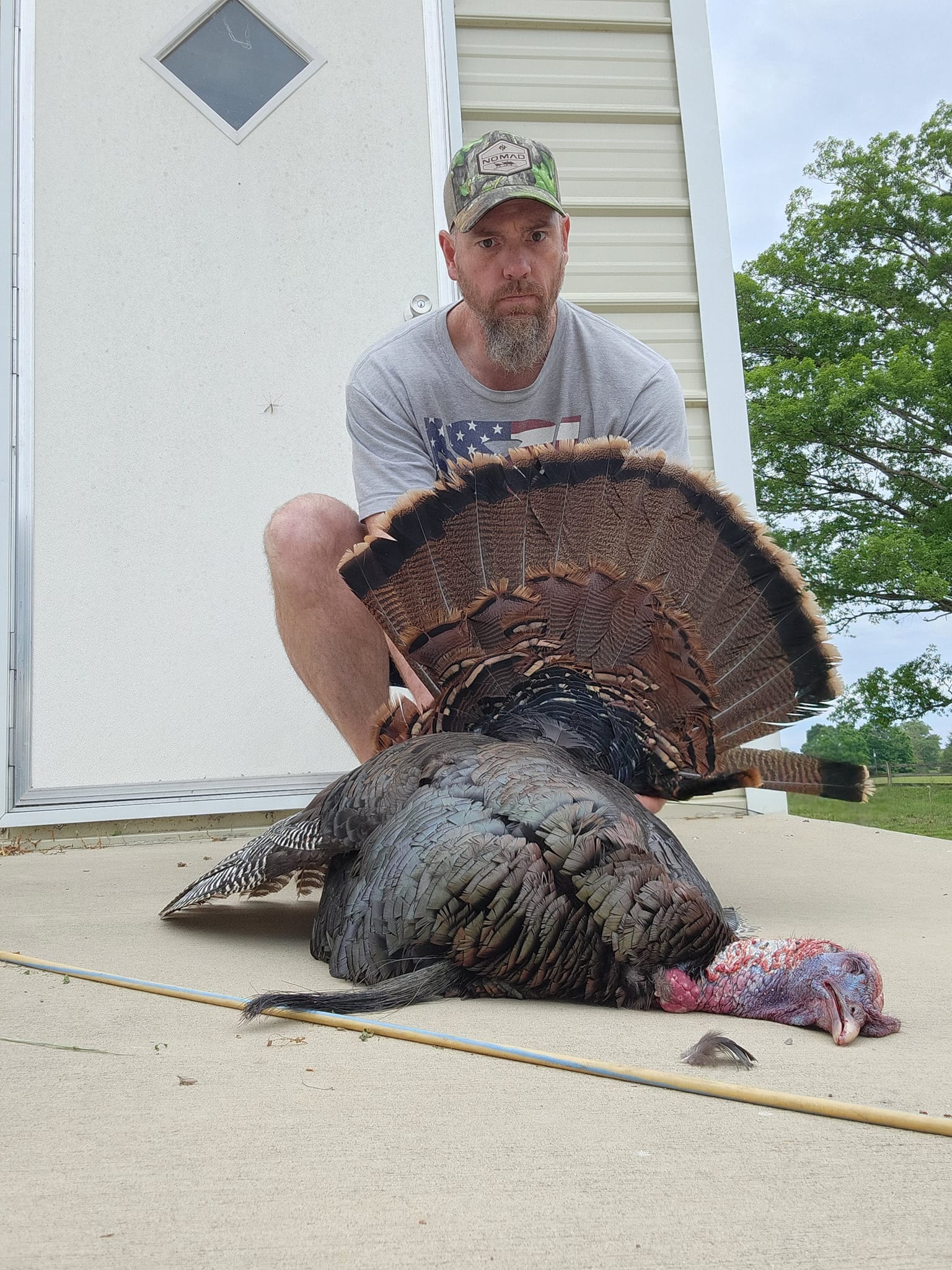 A man kneels behind a harvested wild turkey, which is lying on the ground.