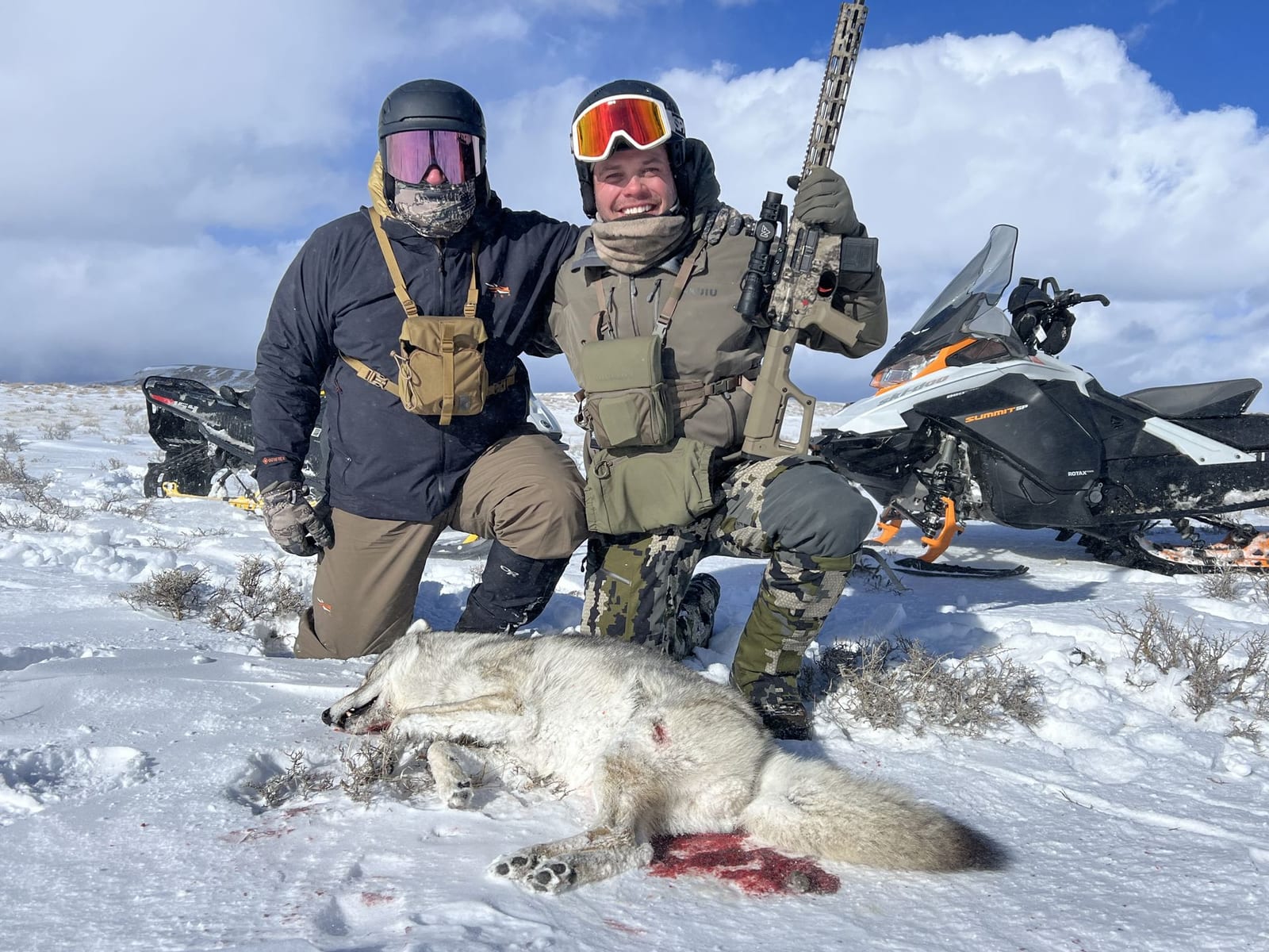 Two hunters pose with a harvested gray wolf in a snowy landscape with snowmobiles in the background.