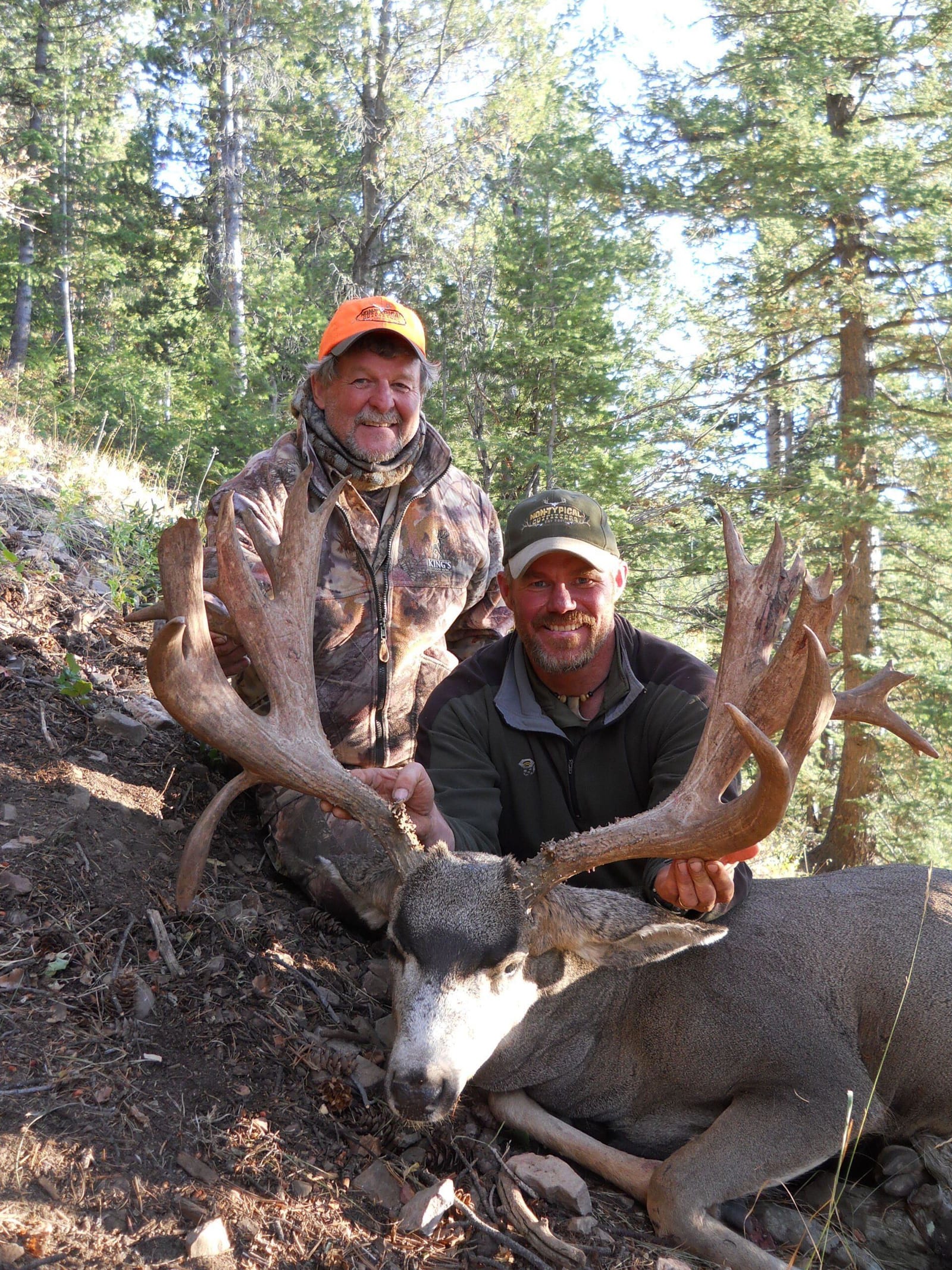 Two men pose with a large buck with impressive antlers in a forested setting.