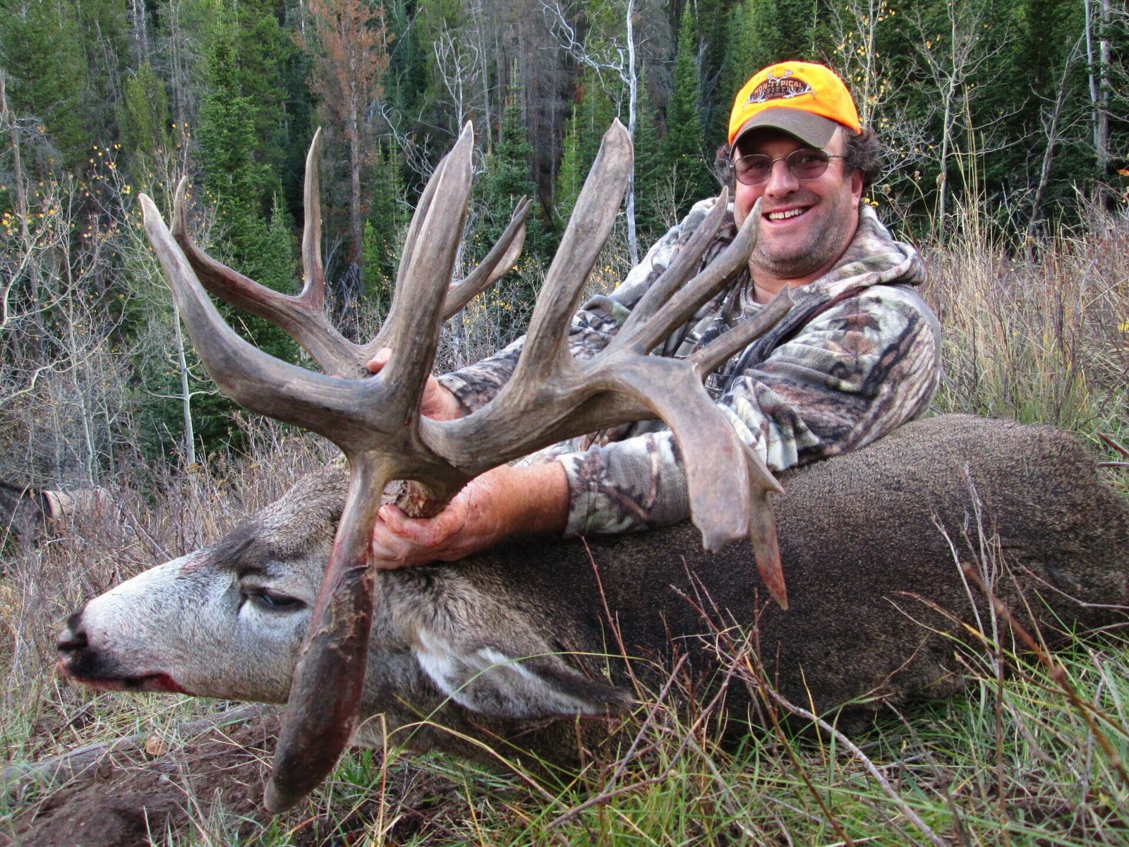 A hunter in camouflage and an orange hat poses with a harvested mule deer.