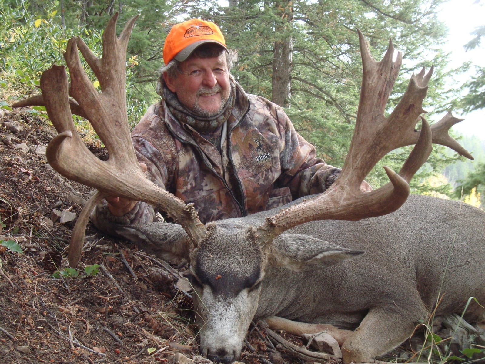 Hunter in camouflage, orange hat, smiling, kneeling next to a large-antlered deer in a forest setting.