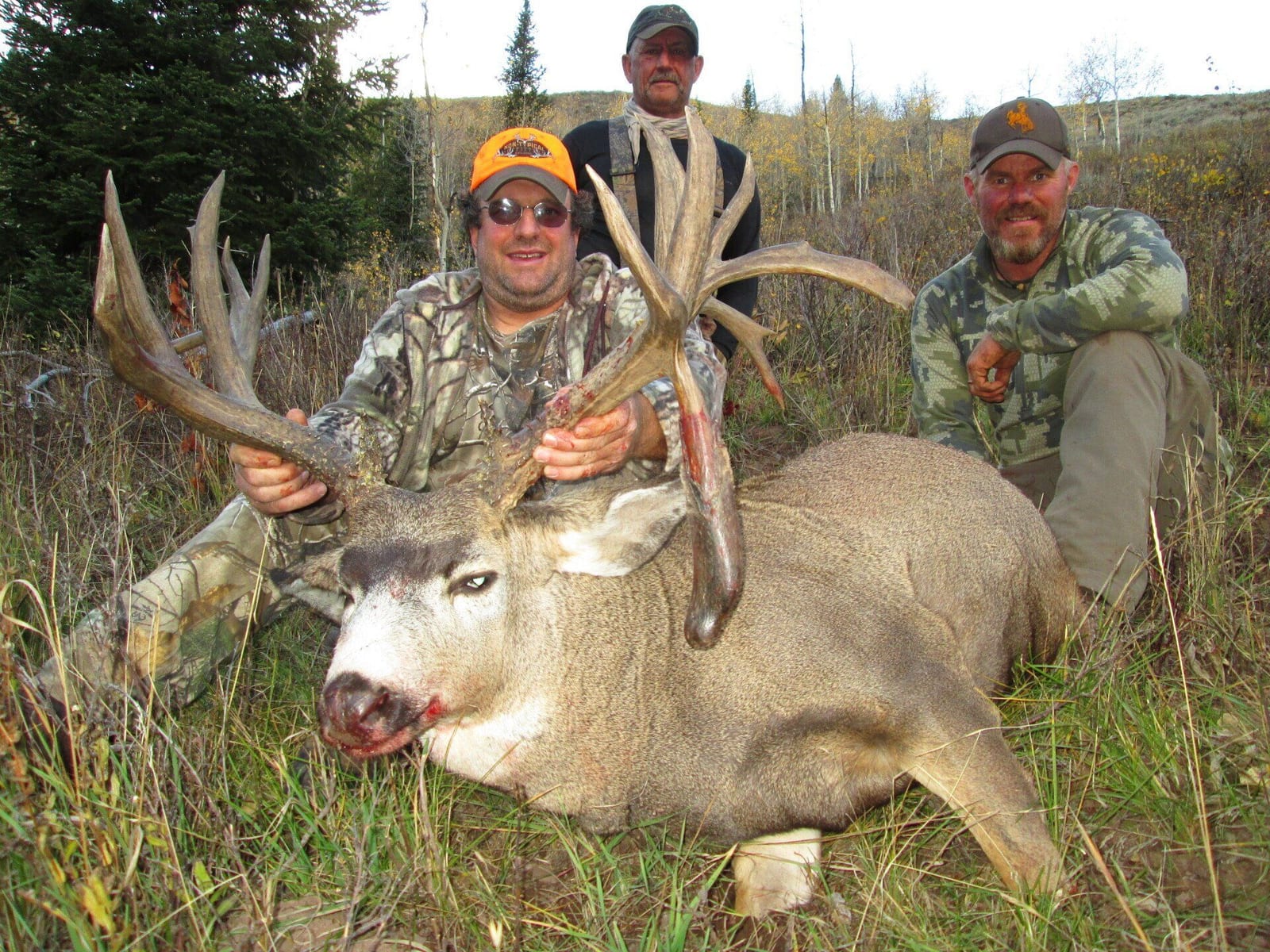 Three hunters pose with a harvested mule deer in a grassy, wooded area.