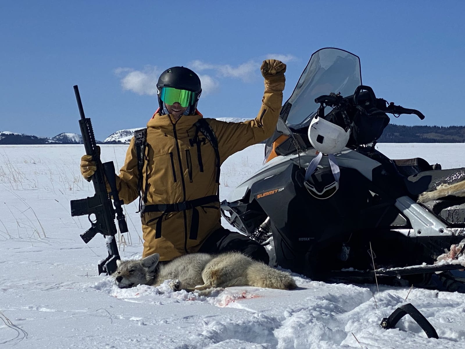 A hunter in winter gear poses with a harvested coyote and a rifle next to a snowmobile in a snowy landscape.