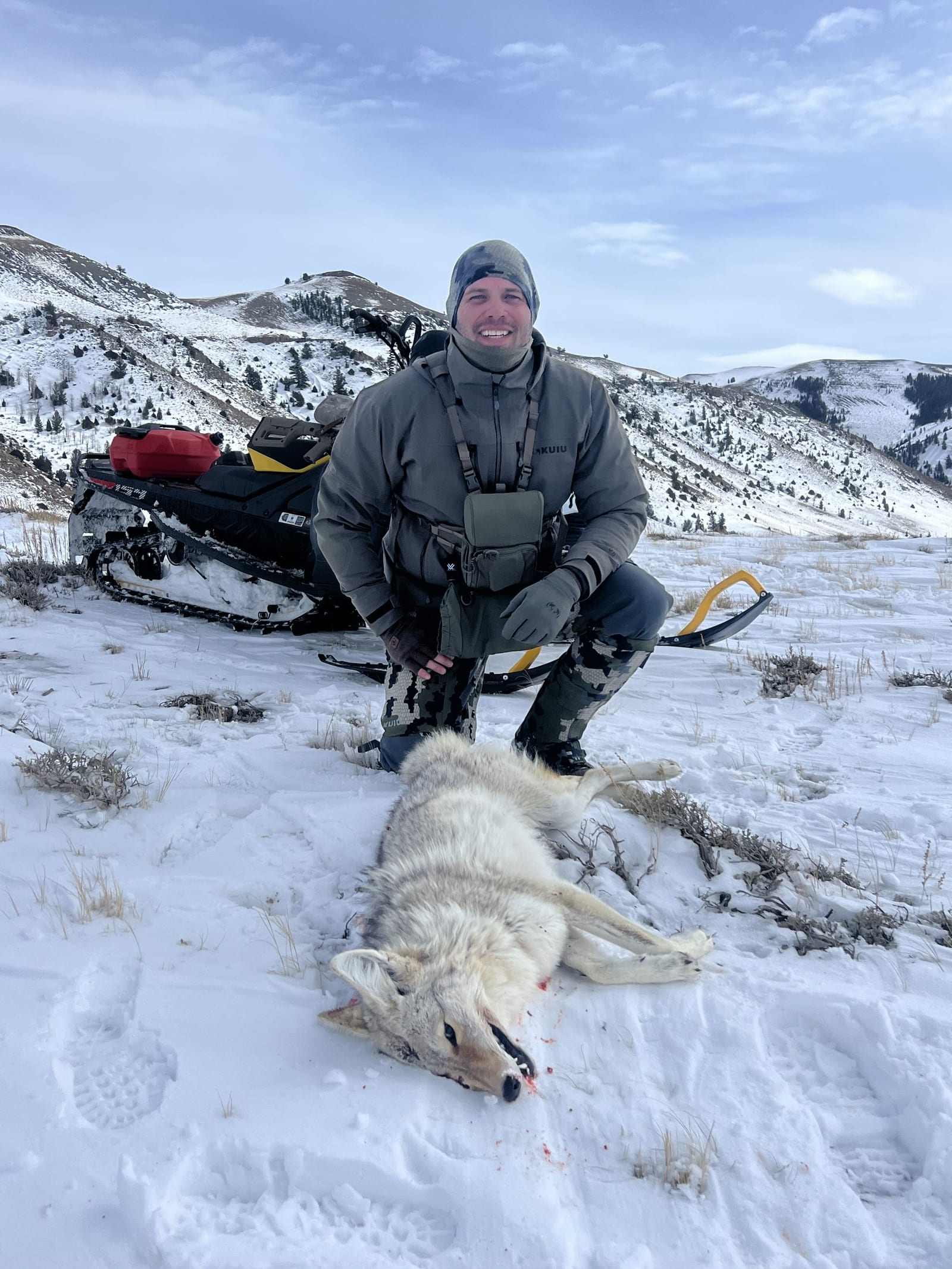 A hunter kneels next to a harvested coyote in a snowy, mountainous landscape with a snowmobile in the background.