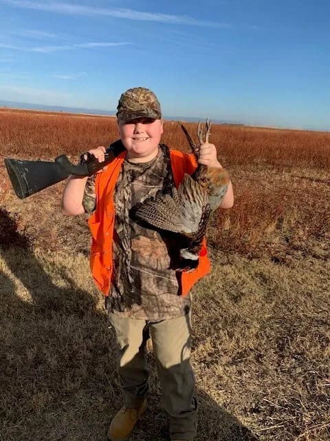 A young hunter in an orange vest and camouflage hat holds a harvested ring-necked pheasant and a shotgun in a field.