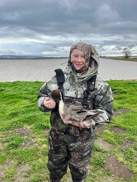 A person in camouflage holds a harvested duck by a body of water under a cloudy sky.