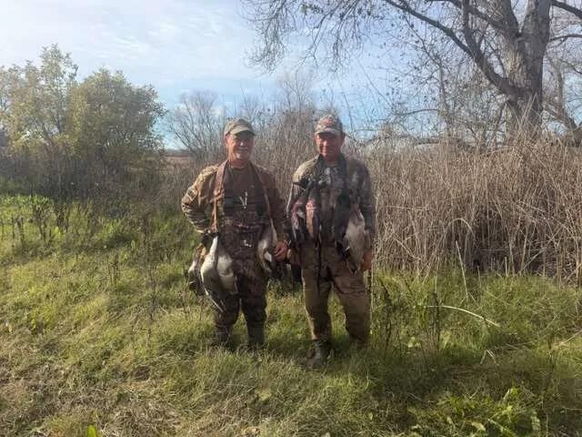 Two hunters in camouflage pose with harvested ducks in a grassy field.