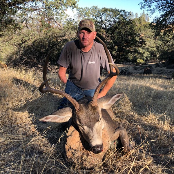 A hunter kneels behind a harvested Columbian blacktail deer in a grassy field.