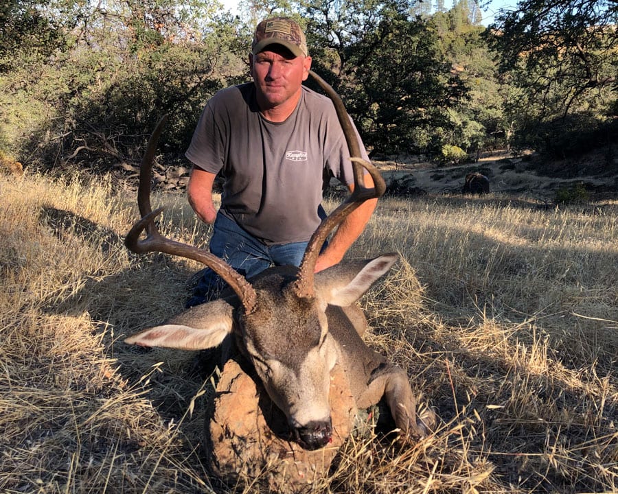 A hunter kneels with a harvested Columbian blacktail deer in a grassy, wooded area.