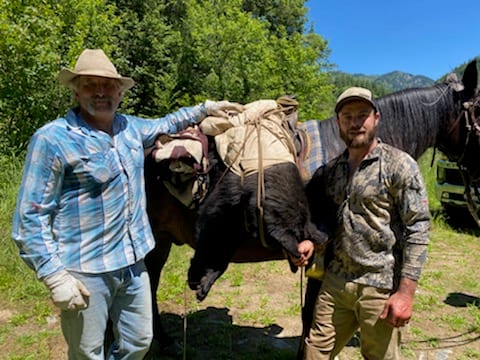 Two men stand next to a horse packed with a harvested black bear in a wooded area.