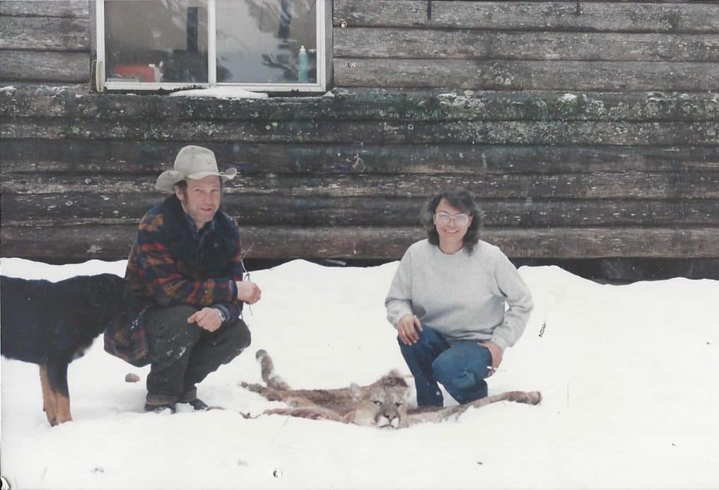 A man and a woman pose with a harvested mountain lion in the snow, with a dog nearby and a wooden building in the background.