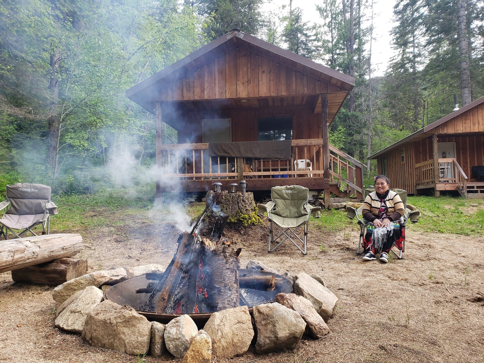 A person sits by a campfire in front of wooden cabins in a forest.