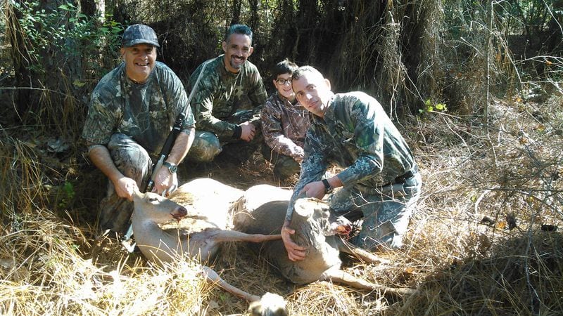 Multiple hunters pose with a harvested whitetail deer in a wooded area.