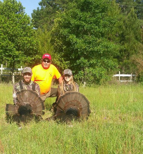 Three hunters pose with two harvested wild turkeys in a grassy field.