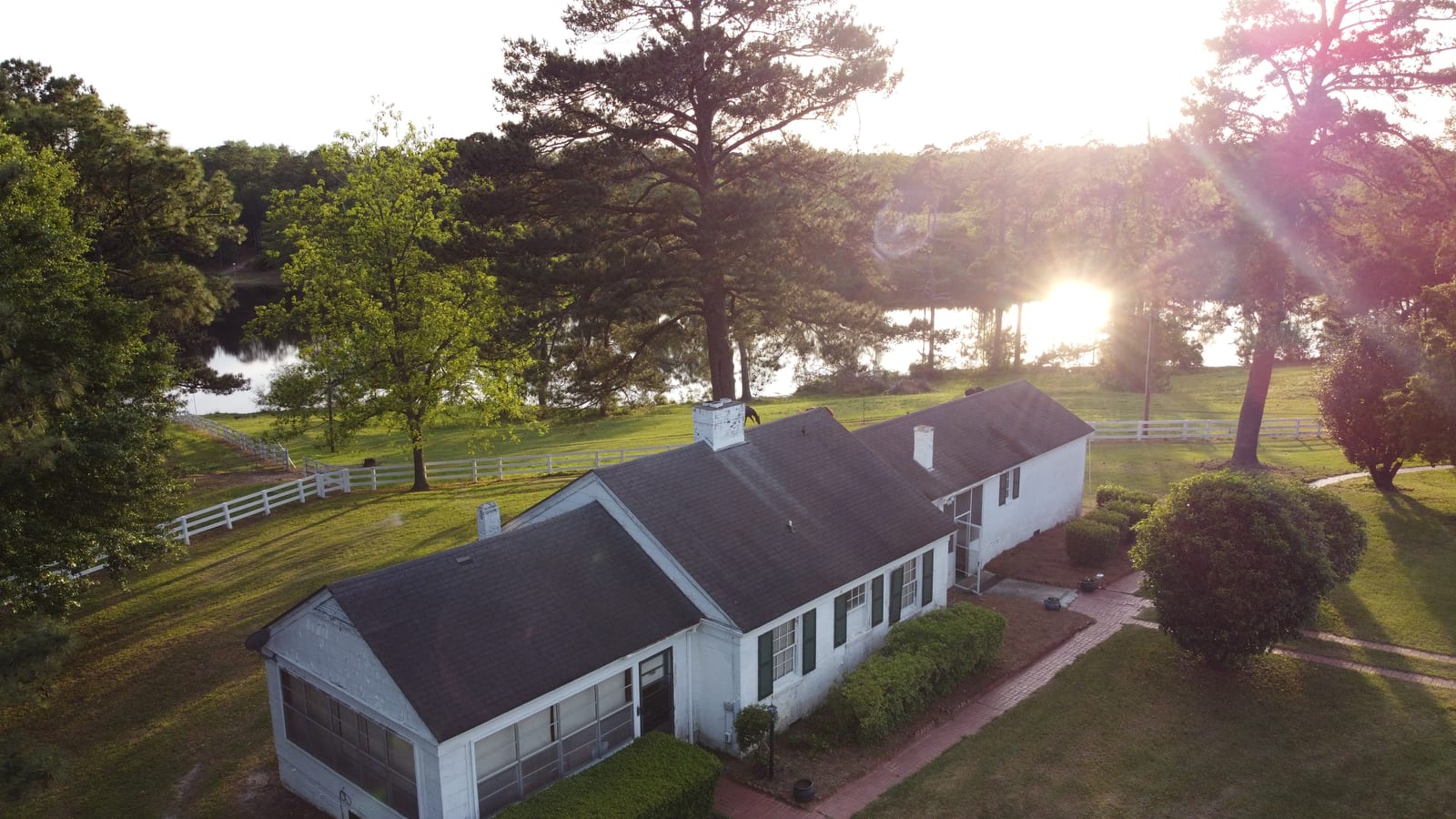 An aerial view of a white lodge building with a dark roof, surrounded by green grass and trees, with a lake in the background at sunset.