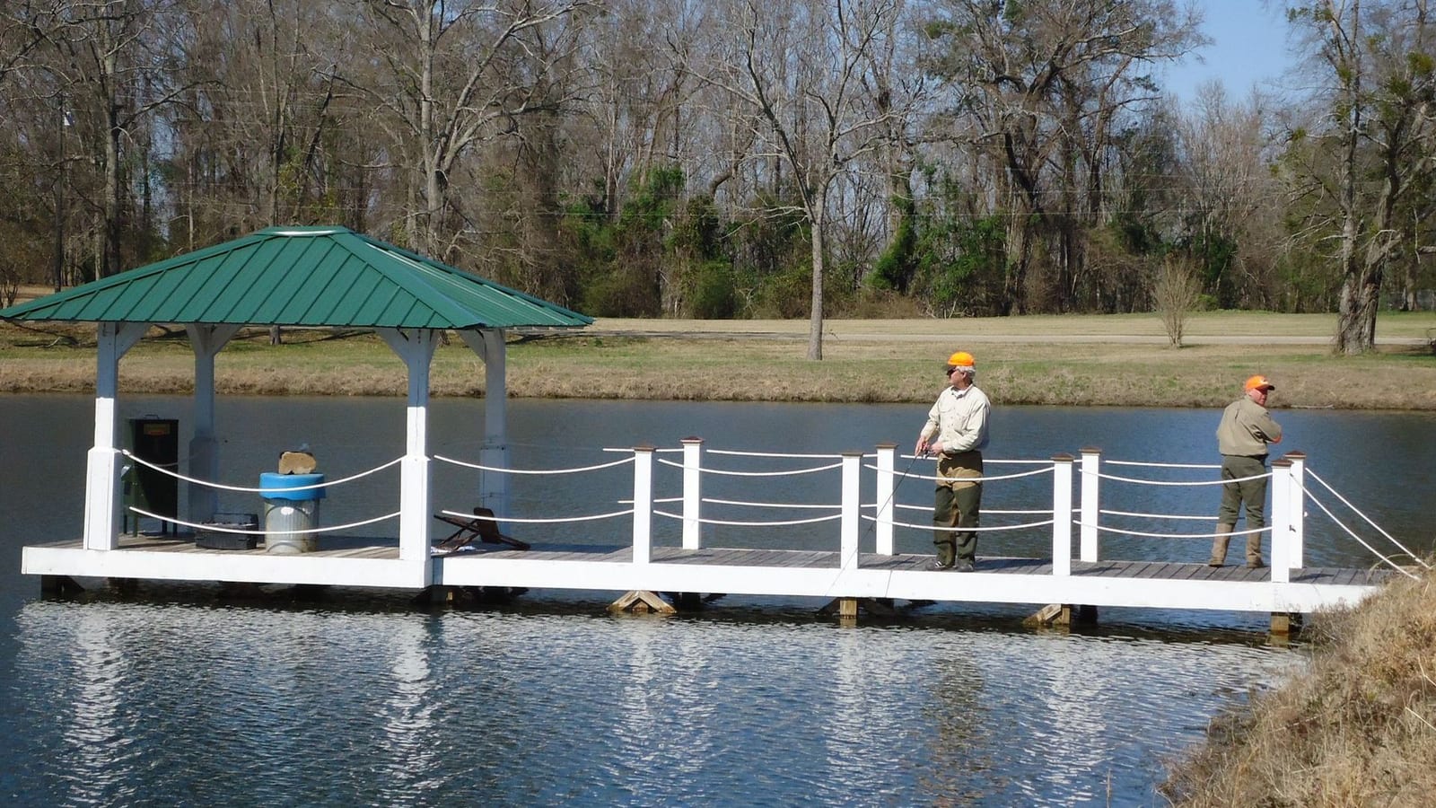Two men stand on a white dock with a green-roofed gazebo over a pond, surrounded by trees.