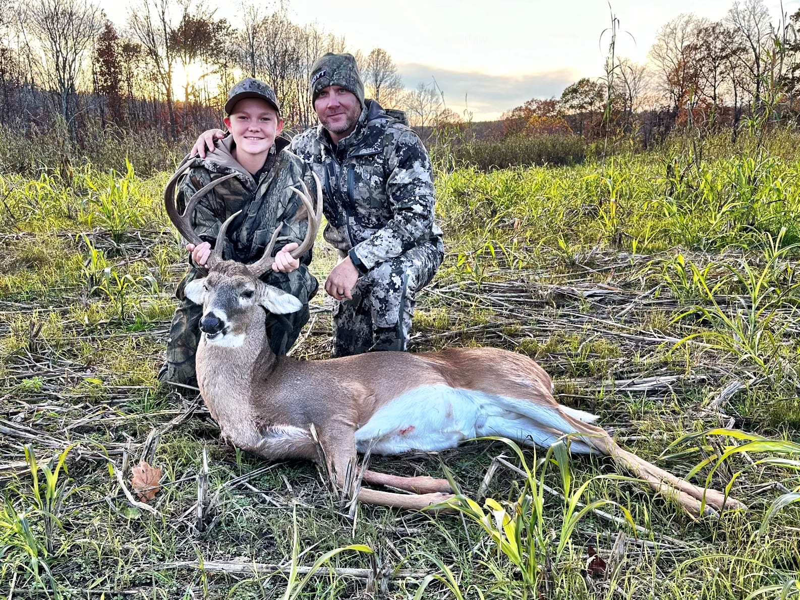 A hunter and a young person pose with a harvested whitetail deer in a grassy field.