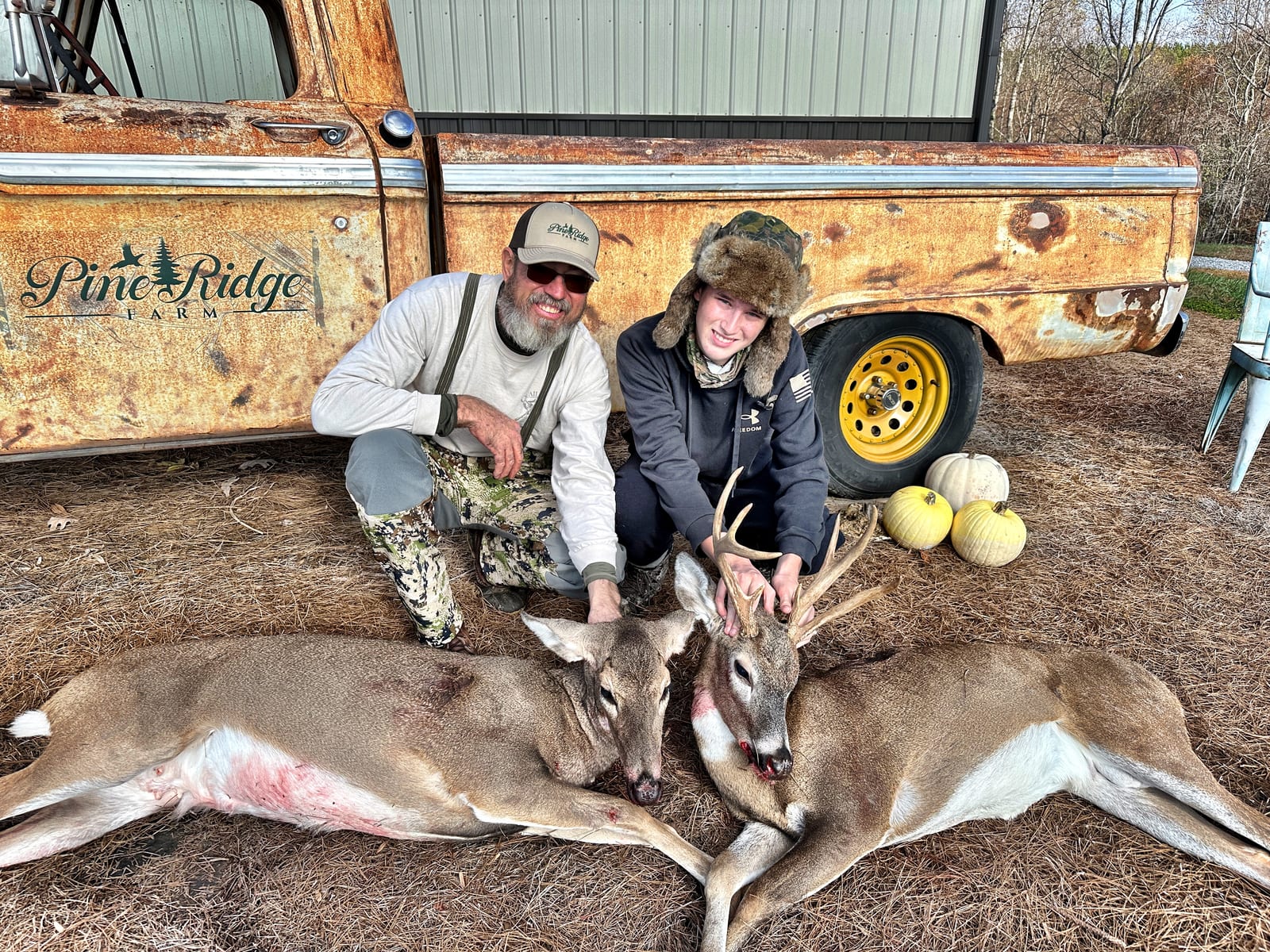 Two hunters pose with two harvested whitetail deer in front of an old truck.